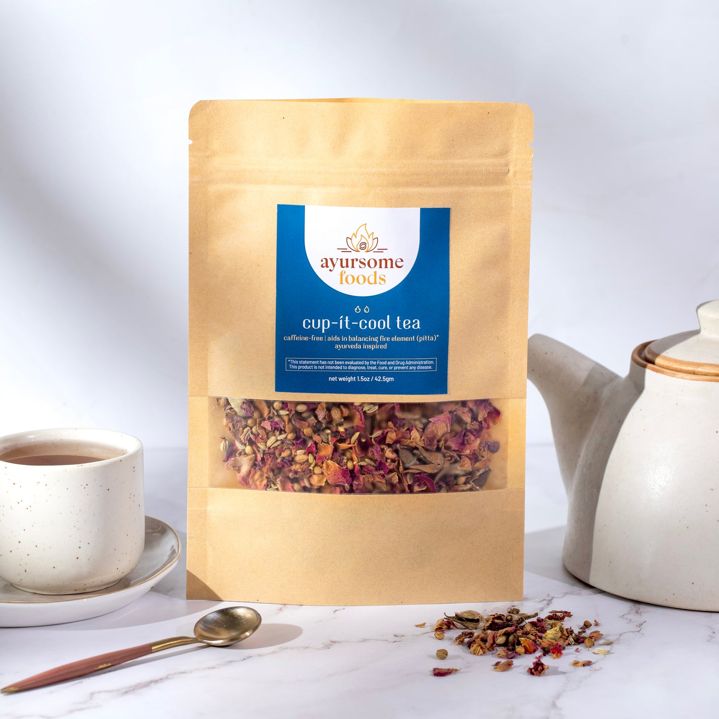 Ayurveda tea for Pitta Dosha that is great for acidity and relaxation. This herbal loose leaf tea is made with organic rose petals and is packed in a recyclable pack. The background is white and next to the loose leaf rose tea is a white kettle, organic tea ingredients, a cup of the brewed tea and a spoon.