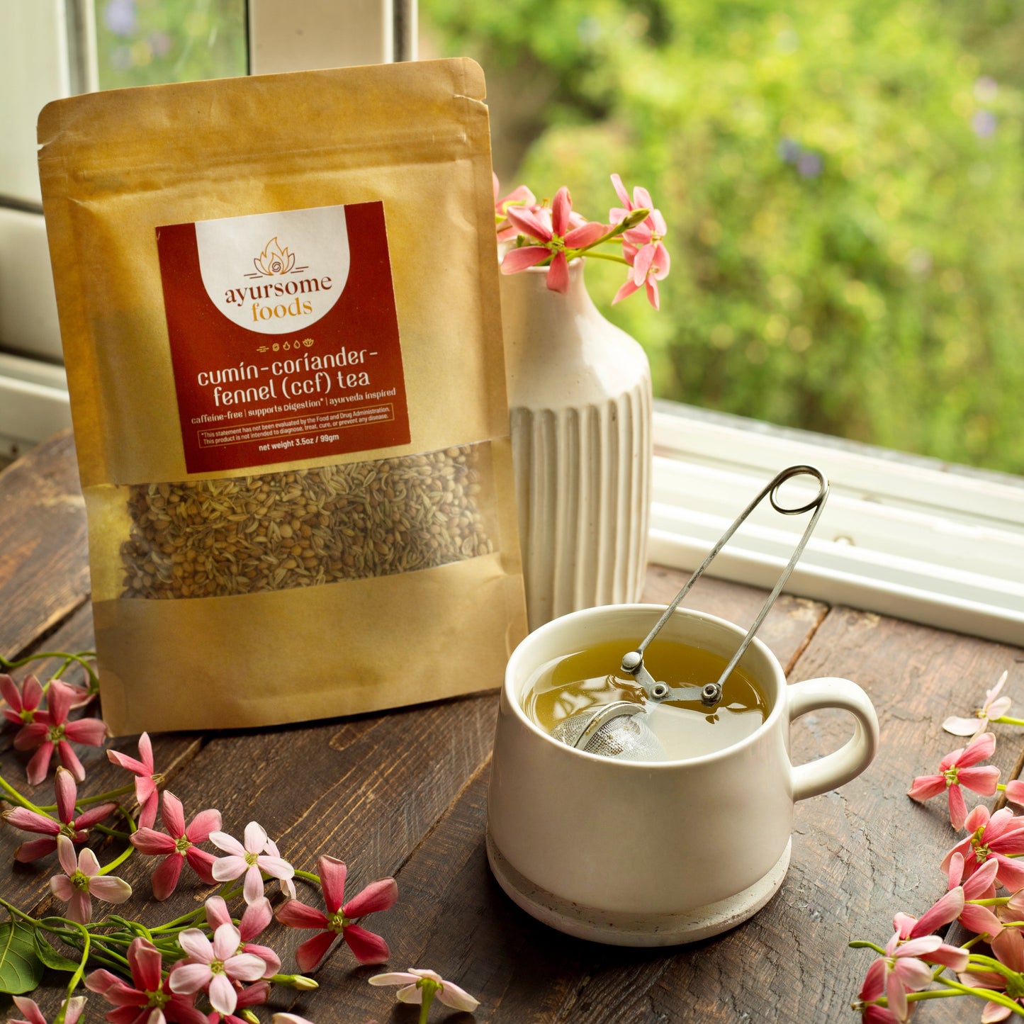 Ayurvedic CCF Tea packed in recyclable brown Kraft paper bag, with a white cup of brewed CCF tea and a tea infuser in the cup, a small white vase with pink flowers and some pink flowers on the window brown sill.