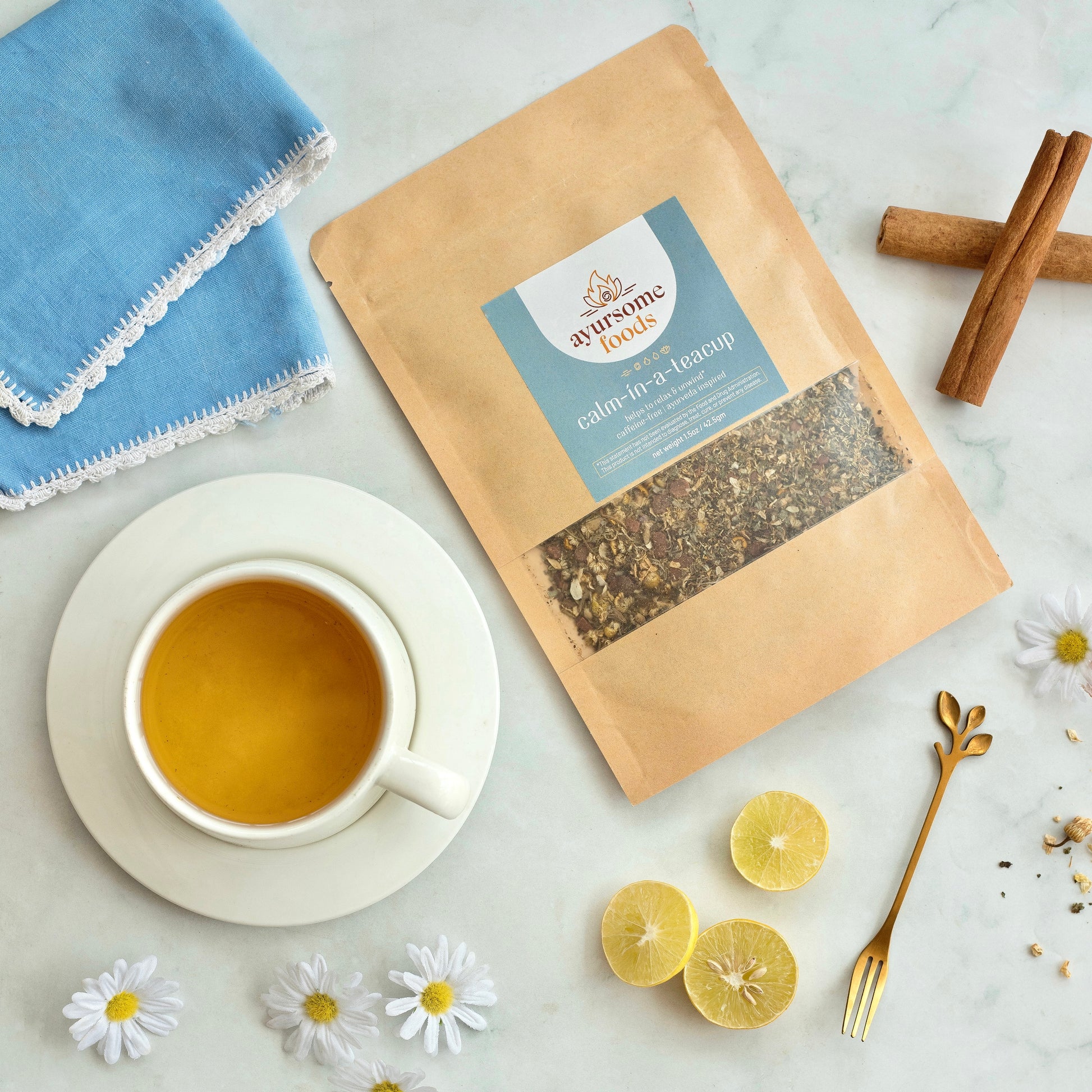 On a white marble background, a pack of the loose leaf chamomile tea for sleep, organic cinnamon sticks, lemon slices, blue napkin, golden fork, a teacup full of herbal night time tea, organic chamomile flowers. This herbal loose leaf lemon balm and chamomile tea is made by Ayursome Foods in NJ, USA.