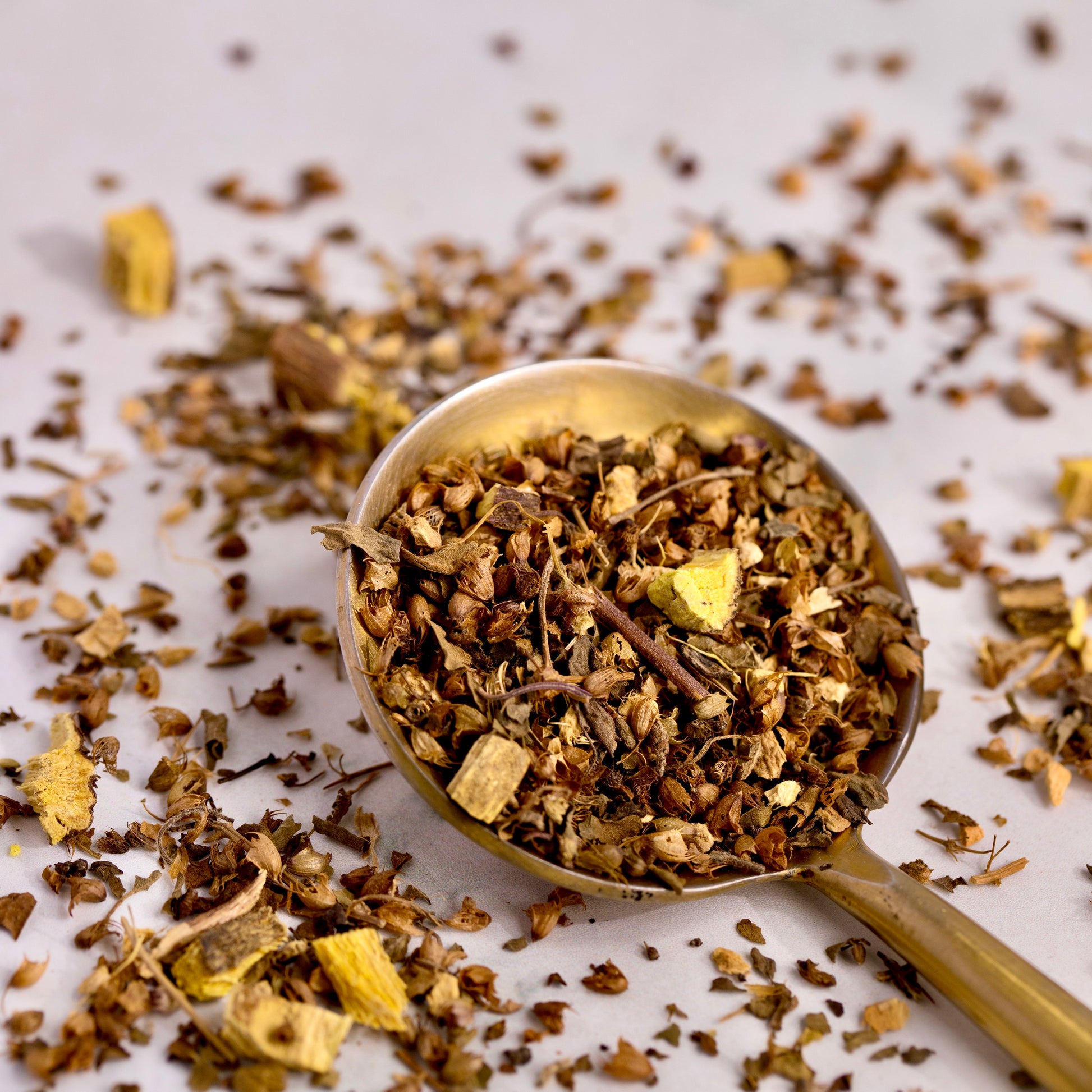 On a white background, there is a gold spoon. In and around the spoon is loose leaf ayurveda tulsi tea blend for cold and cough. The tea blend consists of organic tulsi, organic ginger and organic licorice root. The herbal tea is green and brown in color.